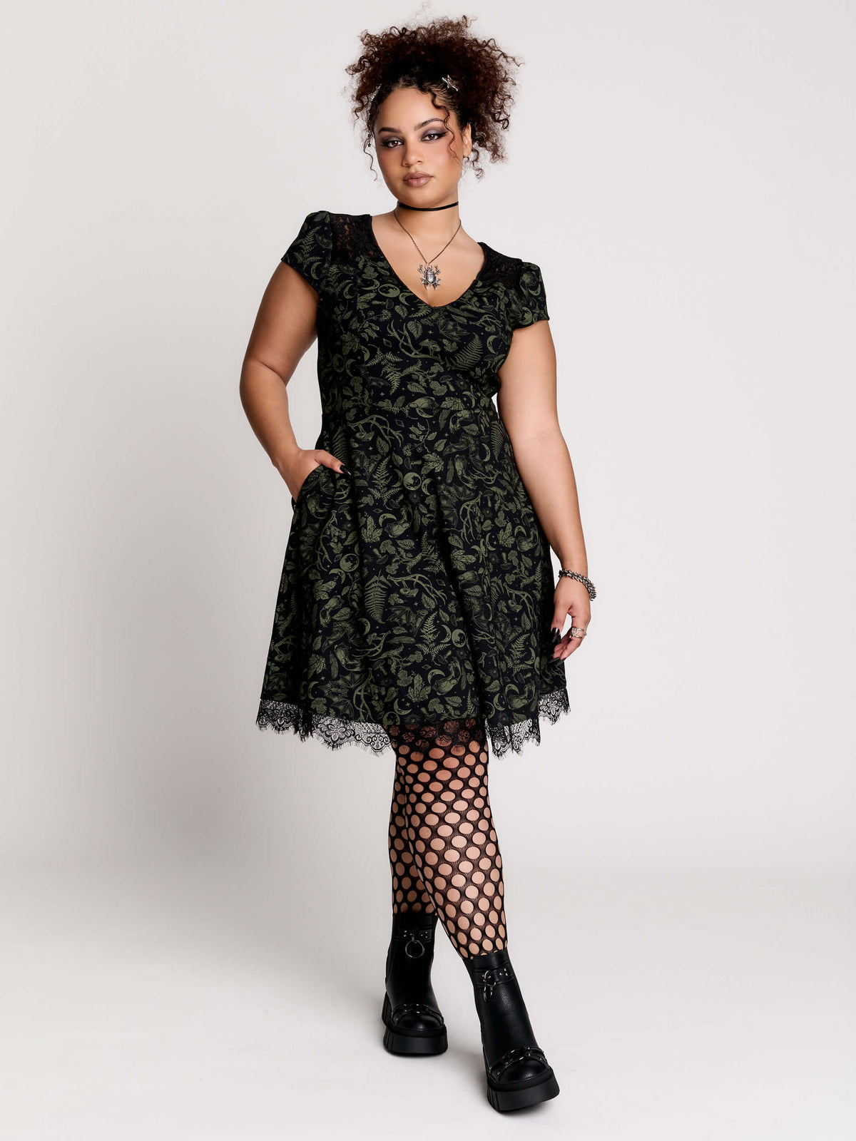 short black dress with a green leafy lace pattern, featuring cap sleeves, a V-neckline, side pockets, and a scalloped lace hem.