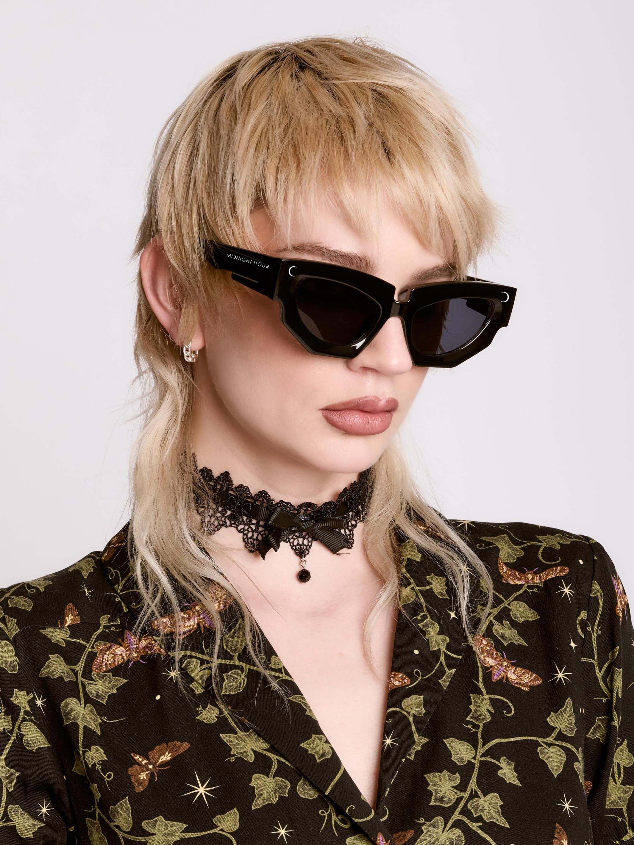 Person wearing black sunglasses and a floral outfit with a choker necklace on a white background.
