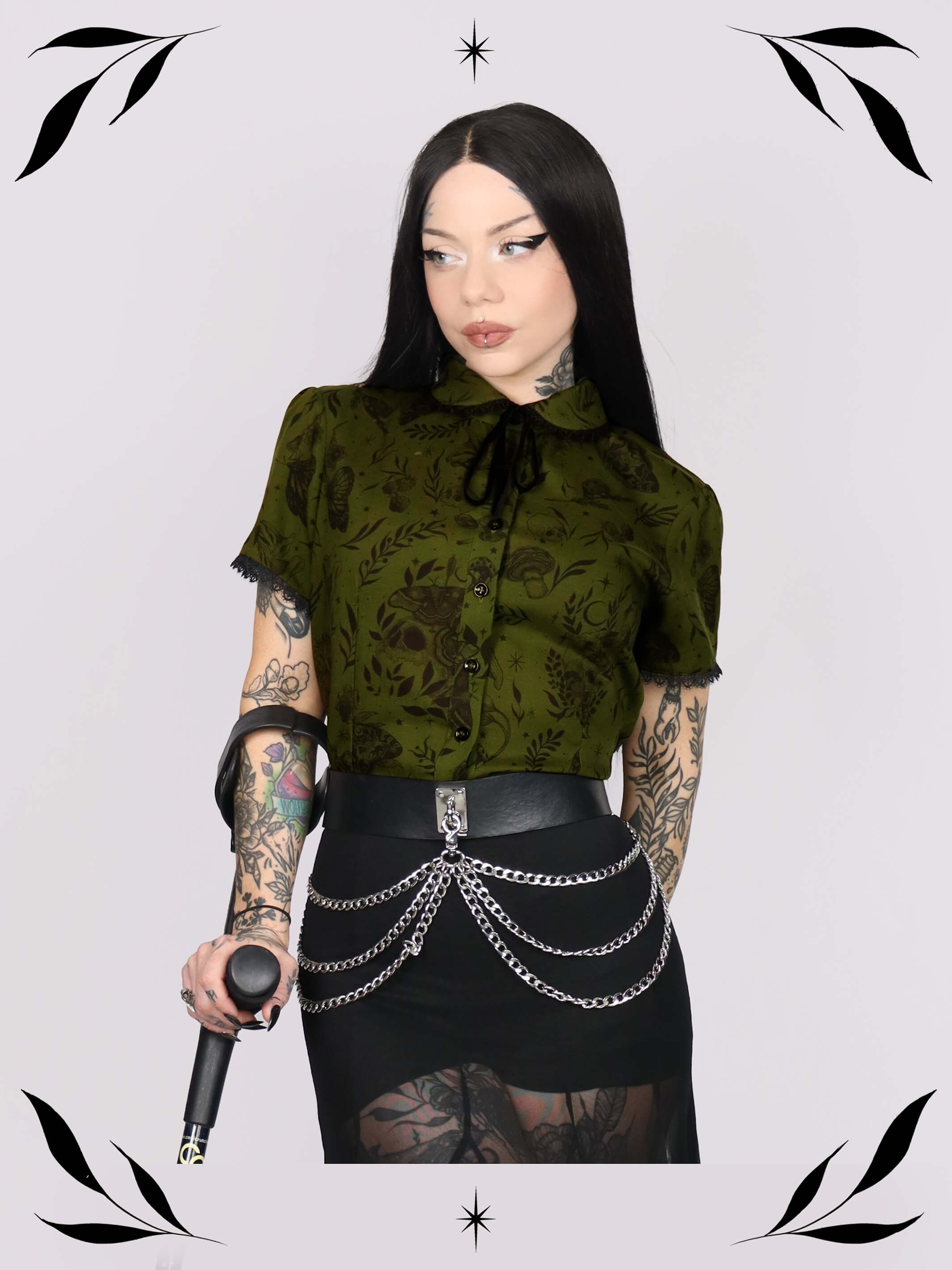 a short-sleeve olive green blouse with a dark botanical pattern, black lace trim collar with black ribbon tiedetail and black buttons.