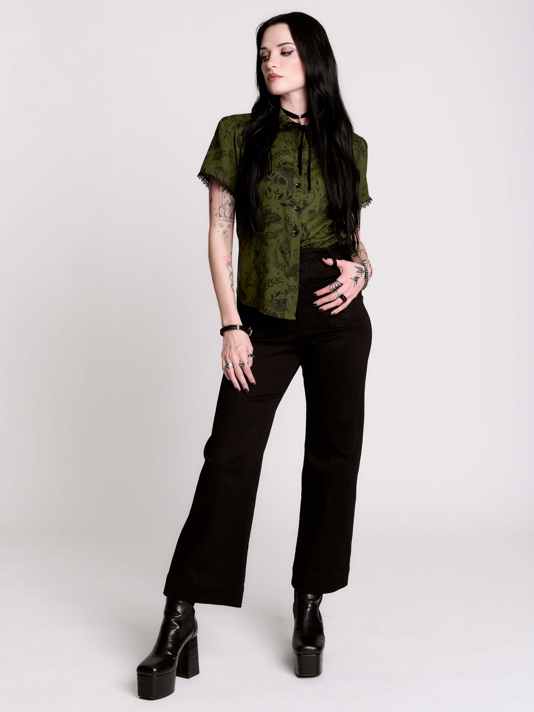 a short-sleeve olive green blouse with a dark botanical pattern, black lace trim collar with black ribbon tiedetail and black buttons.