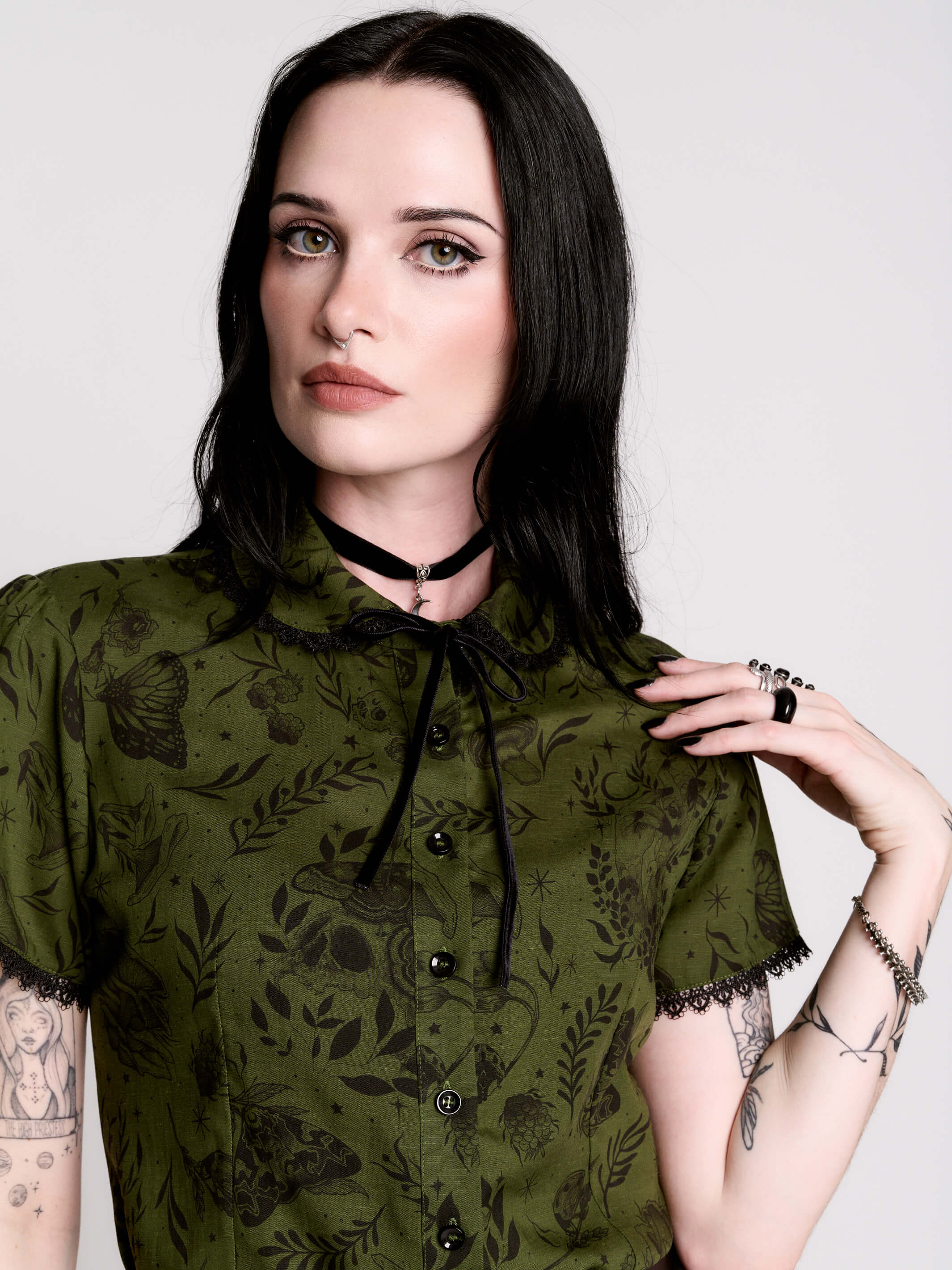 a short-sleeve olive green blouse with a dark botanical pattern, black lace trim collar with black ribbon tiedetail and black buttons.
