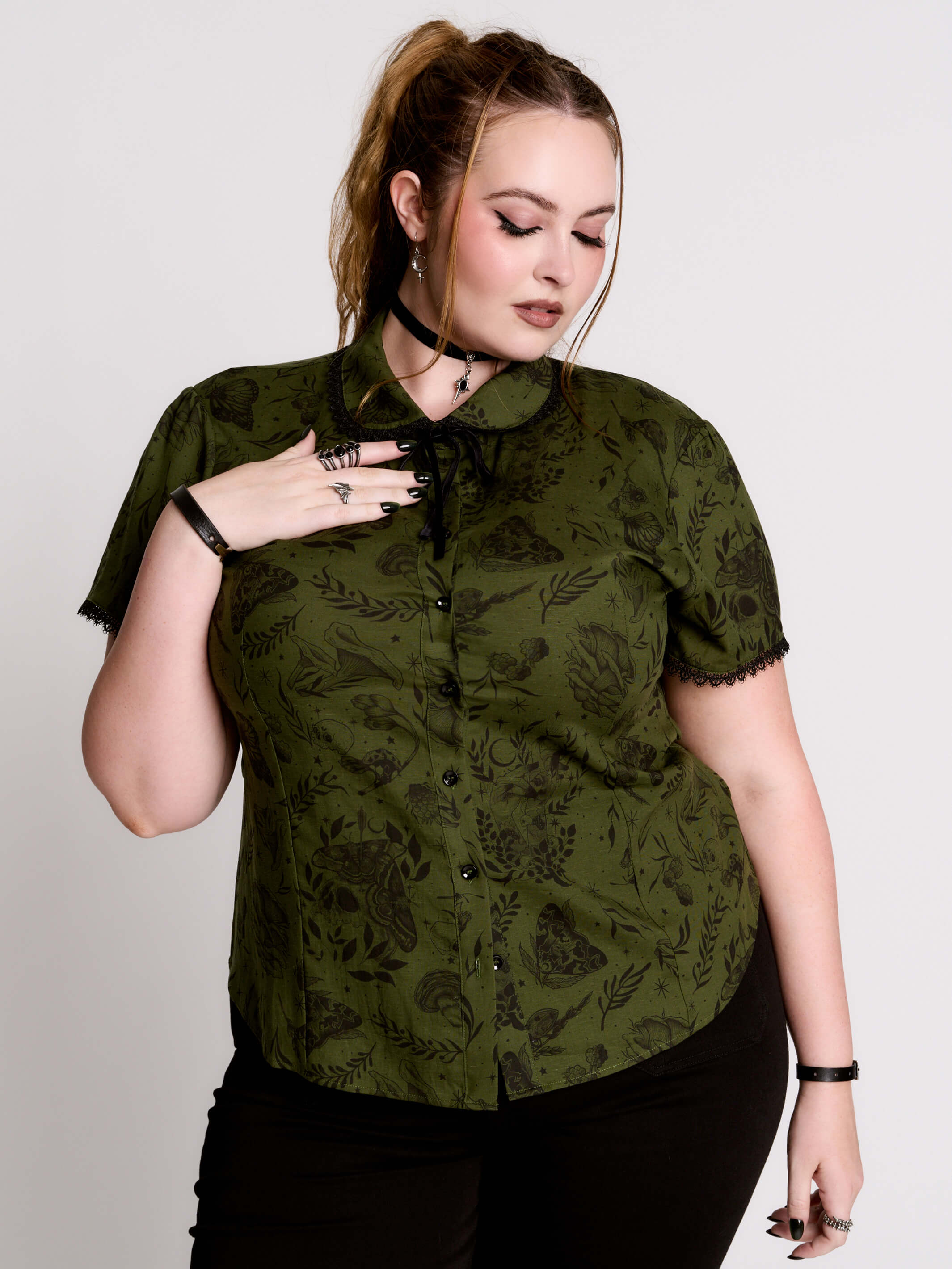 a short-sleeve olive green blouse with a dark botanical pattern, black lace trim collar with black ribbon tiedetail and black buttons.