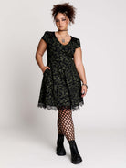 short black dress with a green leafy lace pattern, featuring cap sleeves, a V-neckline, side pockets, and a scalloped lace hem.