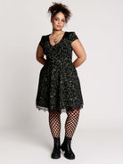 short black dress with a green leafy lace pattern, featuring cap sleeves, a V-neckline, side pockets, and a scalloped lace hem.